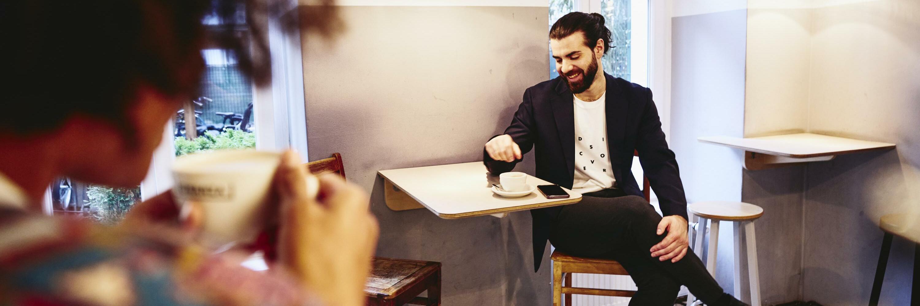 Man sitting at a table with coffee, looking down while having a conversation with someone.