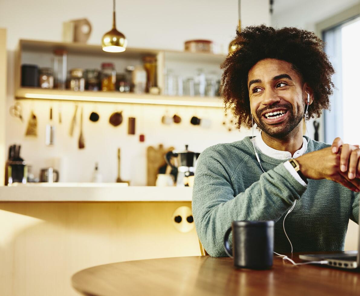 Smiling man sitting at his dining table with a drink and his laptop. Looking away. Kitchen in the background.