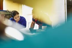 Two blue-collar workers deliberating. Female Asian and male Caucasian. Smiling. Ponytail. Groomed beard. Primary color blue. Secondary color cream/white.