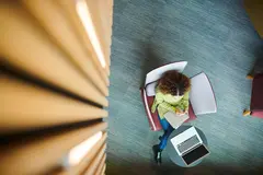 Aerial view of woman writing in a notebook with a computer next to her.